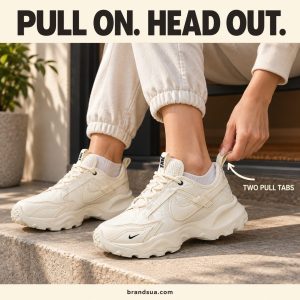 Nike Women's TC 7900 Sneakers DD9682-100 — Seated cropped adult hand naturally grasps rear cream pull loop to guide heel int…